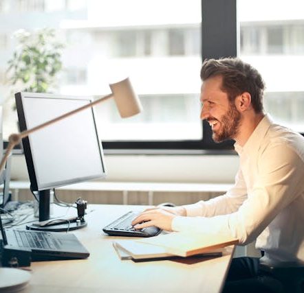 Man in White Dress Shirt Sitting on Black Rolling Chair While Facing Black Computer Set and Smiling
